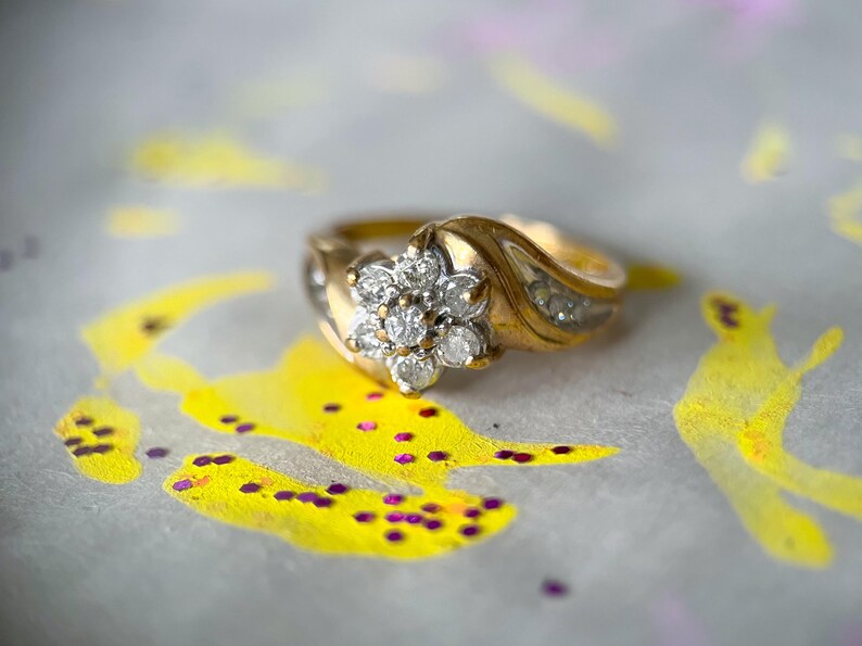 May include: A gold ring with a floral cluster of diamonds at the center. The band features a wave design with smaller diamonds. The ring is set against a background with yellow and purple paint.