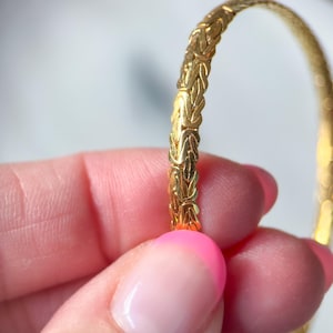 May include: A gold-colored bracelet with a detailed, woven design. The bracelet has a textured surface and a rounded shape. The intricate pattern gives the bracelet a classic and elegant look. The bracelet is held in a person's hand.