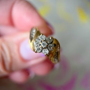 May include: A gold ring featuring a cluster of small, clear gemstones arranged in a flower shape. The ring has a curved band with additional small gemstones. The ring is held up against a soft, blurred background.