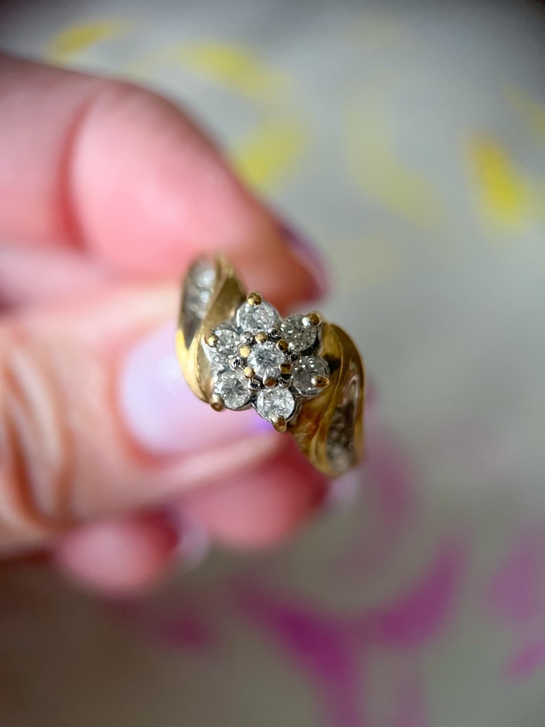 May include: A gold ring featuring a cluster of small, clear gemstones arranged in a flower shape. The ring has a curved band with additional small gemstones. The ring is held up against a soft, blurred background.