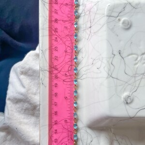 May include: A gold-toned bracelet with oval, light blue gemstones. The bracelet is measured with a pink ruler, showing a length of approximately 7 inches. The ruler is marked with inch increments.