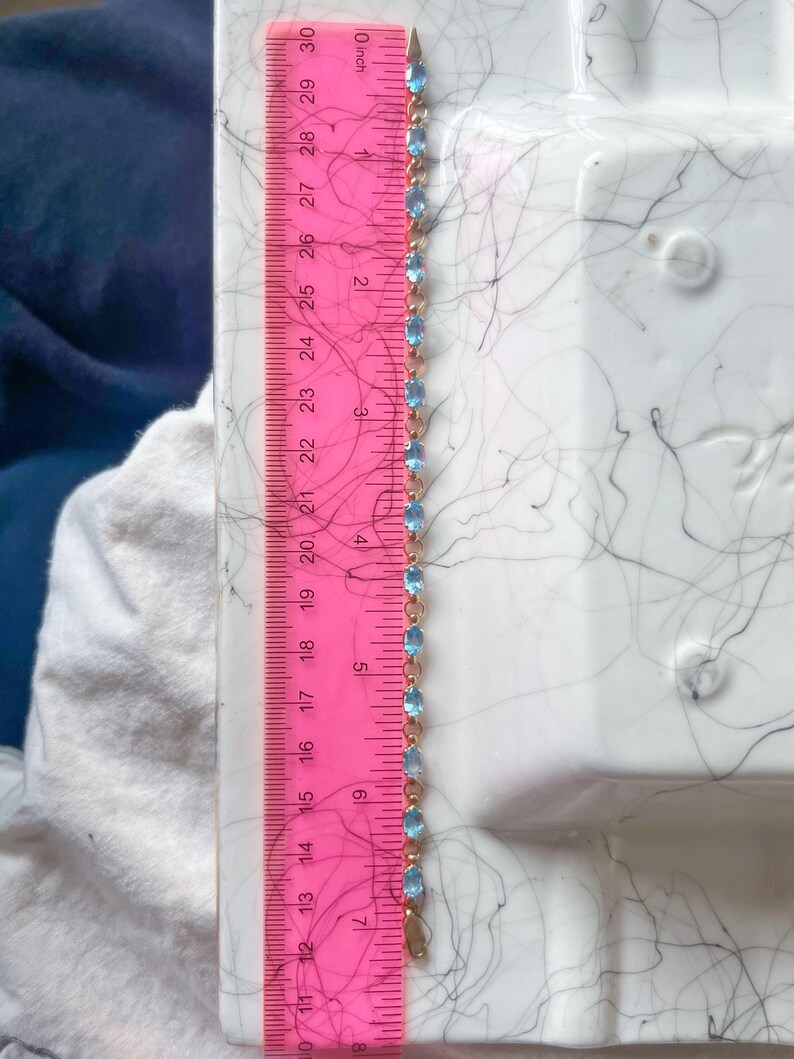 May include: A gold-toned bracelet with oval, light blue gemstones. The bracelet is measured with a pink ruler, showing a length of approximately 7 inches. The ruler is marked with inch increments.