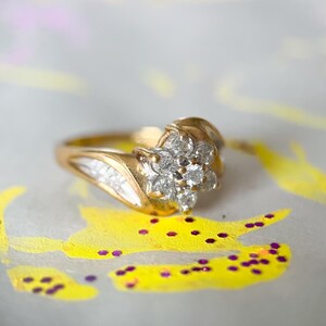 May include: A gold ring with a cluster of small, clear gemstones in a floral arrangement. The ring sits on a white surface with yellow and purple paint details.