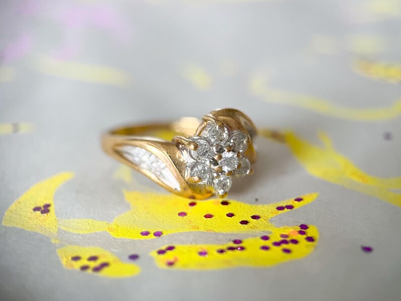 May include: A gold ring with a cluster of small, clear gemstones in a floral arrangement. The ring sits on a white surface with yellow and purple paint details.