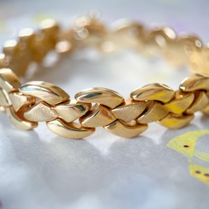May include: A gold-colored bracelet with a linked design. The bracelet is made of multiple, interlocking, leaf-shaped segments. The jewelry is displayed on a patterned surface.
