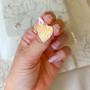May include: A gold heart-shaped pendant with a sunburst design. The pendant is held in a hand with a gold ring and pink nail polish. The background is a white surface with gray marbling.