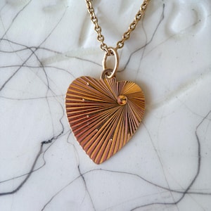 May include: A gold-toned necklace featuring a heart-shaped pendant. The pendant has a sunburst design with a small, round embellishment at the center. The chain is delicate, and the necklace is displayed against a white background.