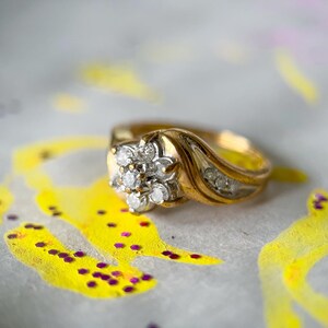 May include: A gold ring with a floral cluster of diamonds at the center. The ring has a curved band with additional small diamonds. The background features yellow and purple paint splatters.