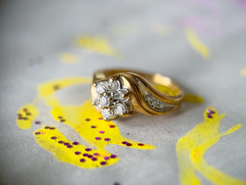 May include: A gold ring with a floral cluster of diamonds at the center. The ring has a curved band with additional small diamonds. The background features yellow and purple paint splatters.