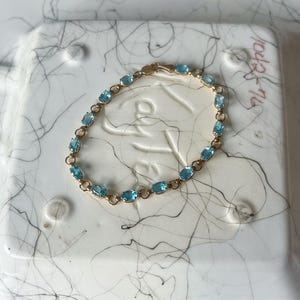 May include: A gold bracelet featuring oval-cut, light blue gemstones. The bracelet has a delicate chain design and a secure clasp. The gemstones are evenly spaced along the bracelet's length, creating a classic and elegant look. This piece of jewelry is suitable for everyday wear or special occasions.