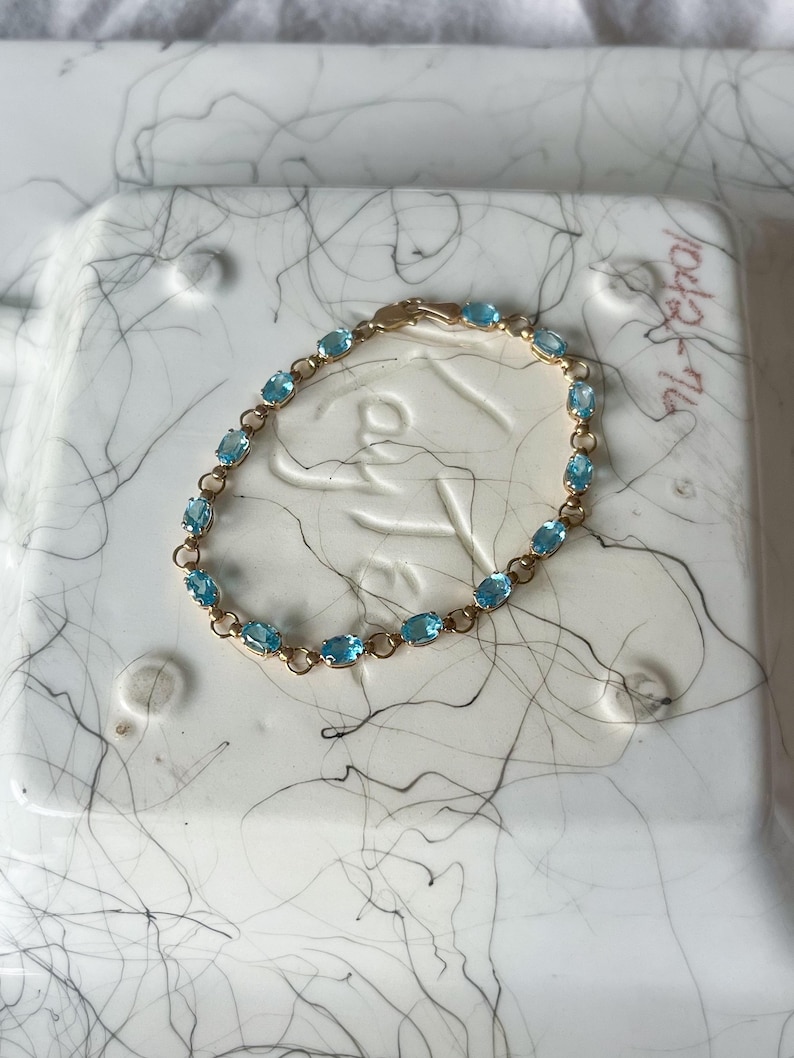 May include: A gold bracelet featuring oval-cut, light blue gemstones. The bracelet has a delicate chain design and a secure clasp. The gemstones are evenly spaced along the bracelet's length, creating a classic and elegant look. This piece of jewelry is suitable for everyday wear or special occasions.