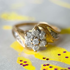 May include: A gold ring with a flower-shaped cluster of diamonds. The ring has a split shank design and is set against a background of yellow and purple paint.