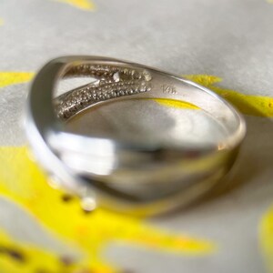 May include: A silver ring with a split band design. One side of the band is encrusted with small, clear gemstones. The ring is marked with "14K". The ring is set against a background with yellow and white accents.