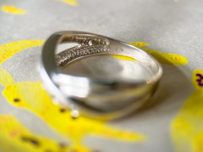 May include: A silver ring with a split band design. One side of the band is encrusted with small, clear gemstones. The ring is marked with "14K". The ring is set against a background with yellow and white accents.