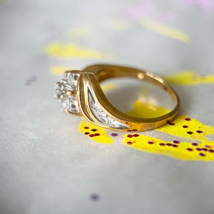 May include: A gold ring with a cluster of small diamonds and a curved design. The ring is set against a background with yellow and purple paint splatters.