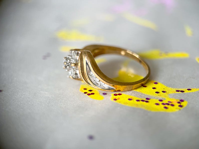 May include: A gold ring with a cluster of small diamonds and a curved design. The ring is set against a background with yellow and purple paint splatters.