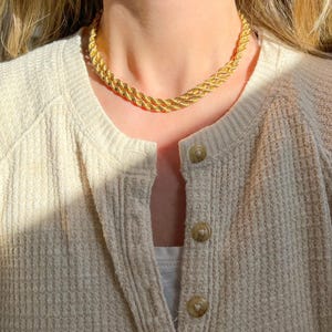 May include: A gold-colored rope chain necklace. The necklace is a choker style, with a twisted rope design. The necklace is worn over a cream-colored cardigan with visible buttons.