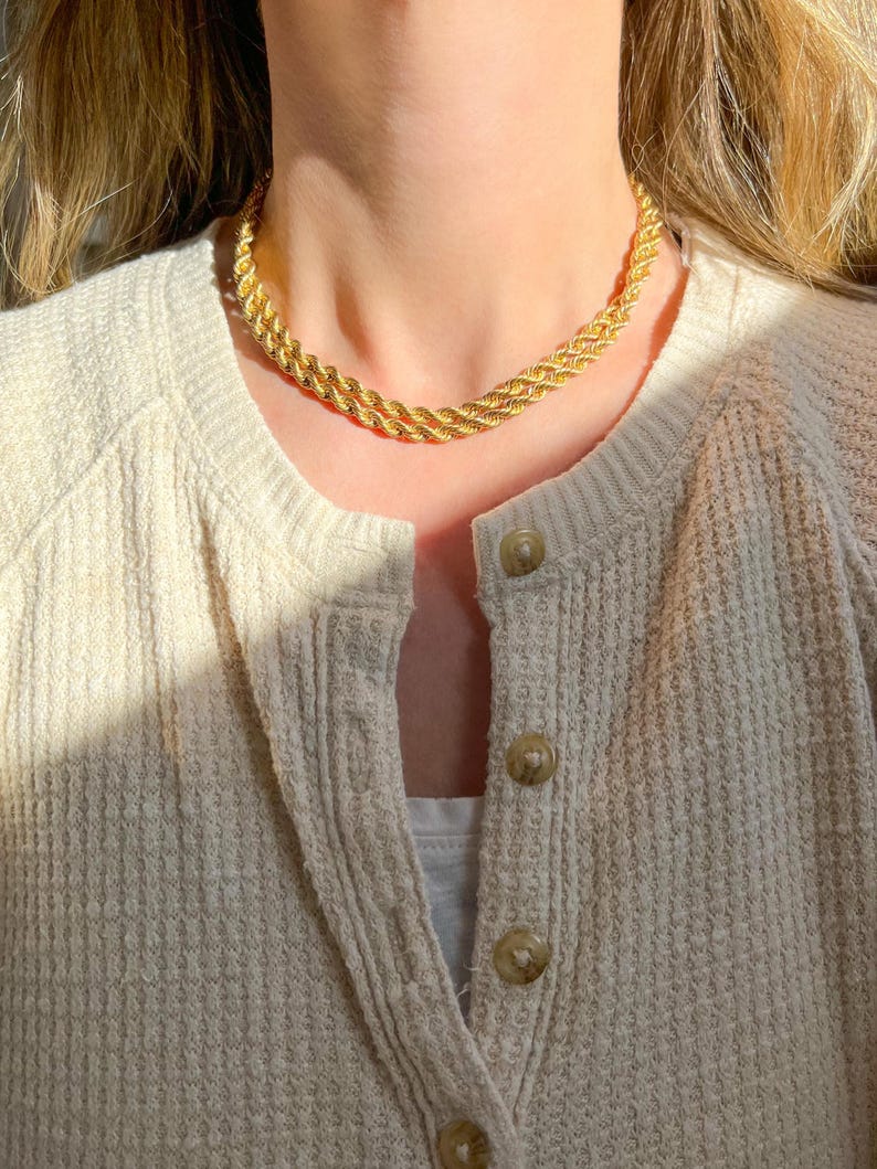 May include: A gold-colored rope chain necklace. The necklace is a choker style, with a twisted rope design. The necklace is worn over a cream-colored cardigan with visible buttons.