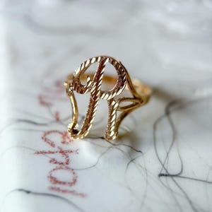 May include: A gold-colored ring with the letter "P" in a script font. The ring is on a white surface with a marble design and red text. The ring's design features a twisted rope-like texture.