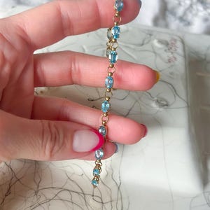 May include: A delicate gold bracelet featuring oval-cut, light blue gemstones. The bracelet is held up against a soft, blurred background. The gemstones are evenly spaced along the gold chain, creating a refined and elegant look.