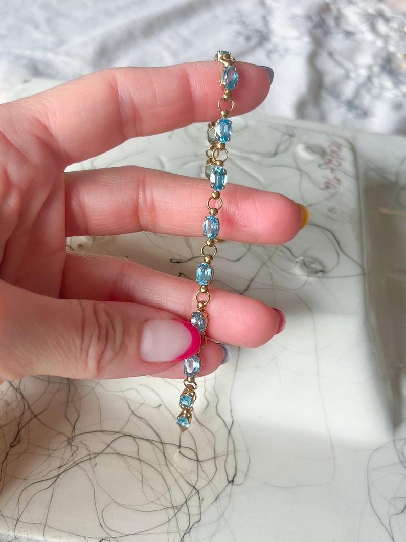 May include: A delicate gold bracelet featuring oval-cut, light blue gemstones. The bracelet is held up against a soft, blurred background. The gemstones are evenly spaced along the gold chain, creating a refined and elegant look.