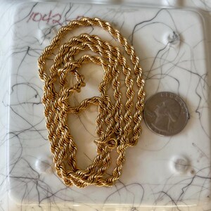 May include: A gold-colored rope chain necklace. The chain is lying on a white surface with black marbling. A U.S. quarter coin is next to the chain for size comparison. The chain is a twisted rope design.