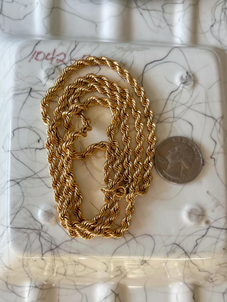 May include: A gold-colored rope chain necklace. The chain is lying on a white surface with black marbling. A U.S. quarter coin is next to the chain for size comparison. The chain is a twisted rope design.