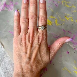 May include: A gold ring with a cluster of small, clear stones is worn on a hand with purple-tipped nails. The ring has a detailed, ornate design. The nails have a gradient effect, transitioning from clear to purple.