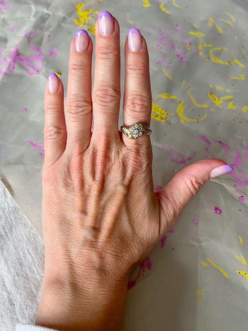 May include: A gold ring with a cluster of small, clear stones is worn on a hand with purple-tipped nails. The ring has a detailed, ornate design. The nails have a gradient effect, transitioning from clear to purple.