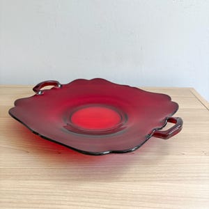 May include: A translucent ruby red glass serving tray with a scalloped edge and two integrated handles on opposite sides. The tray features a smooth, circular center depression and a wide, flat rim, showcasing a vibrant, deep red color throughout.