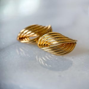 May include: A pair of gold-colored earrings. Each earring features a woven design, with multiple strands of gold wire creating a textured, oval shape. The earrings are displayed on a light-colored surface, reflecting the light and highlighting their metallic sheen.