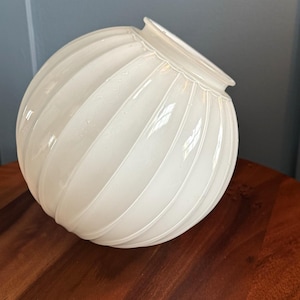 May include: A white, ribbed glass globe with a rounded shape. The globe has a wide opening at the top and is resting on a wooden surface. The ribbed design creates a textured appearance.