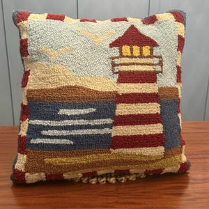 May include: Square decorative pillow with a lighthouse design. The pillow features a red and white striped lighthouse, a blue ocean, and a sandy beach. The pillow has a textured, woven appearance and is trimmed with a red border.