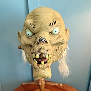 May include: A tan-colored latex mask featuring a wrinkled, aged face with deep-set, pale green eyes, a wide mouth with prominent yellowed teeth, and thin, wispy white hair strands extending from the sides of the head and jawline area.