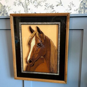 May include: Framed artwork featuring a detailed portrait of a horse's head. The horse has a brown coat with a white blaze and flowing blonde mane. The artwork is set against a light orange background, framed with a black mat and a wooden frame.