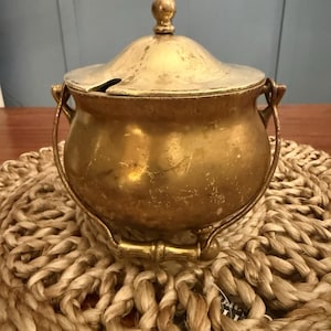 May include: A vintage brass sugar bowl with a lid and handle. The bowl has a rounded shape with a small handle on each side and a lid with a small knob on top. The brass has a tarnished appearance, giving it a rustic look. The bowl sits on a woven placemat.