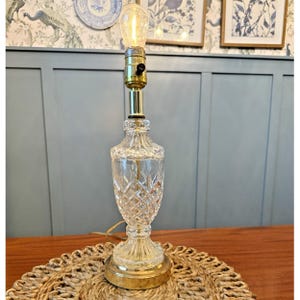 May include: A clear glass table lamp with a diamond pattern on the body, a brass base, and a brass neck. The lamp has a vintage-style lightbulb. The lamp is sitting on a woven mat.
