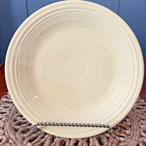 May include: A pale yellow ceramic plate with concentric rings near the rim. The plate is displayed on a silver-colored metal plate stand, resting on a woven placemat. The plate has a smooth, glossy finish.
