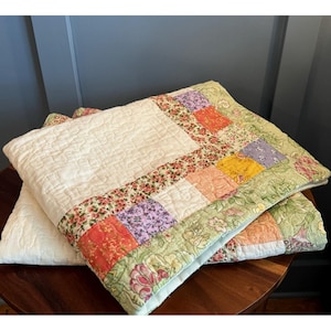 May include: Two handmade quilts with a cream-colored center and floral patchwork borders. The borders feature a variety of colors, including red, orange, purple, and green. The quilts are folded and stacked on a wooden surface.