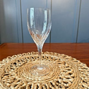 May include: A clear glass wine glass with a delicate stem and a wide bowl. The glass is sitting on a woven placemat.