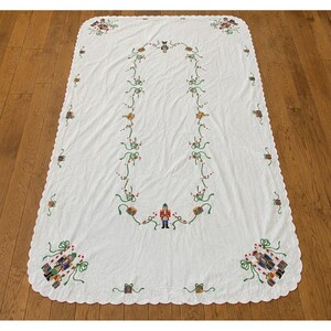 May include: A white rectangular tablecloth with a scalloped edge, featuring embroidered Christmas motifs including nutcrackers, wrapped gifts, green ribbons, and red berries arranged in a central oval pattern and at the corners.