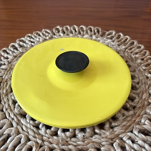 May include: A bright yellow lid with a black knob. The lid is round and appears to be made of enameled metal. The lid is sitting on a woven, circular placemat. The knob is a small, circular shape.