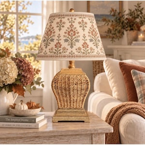May include: A table lamp with a woven natural fiber base and a tapered fabric lampshade featuring a repeating floral pattern in muted red, green, and beige tones. The lamp sits on a square wooden base and provides soft illumination.