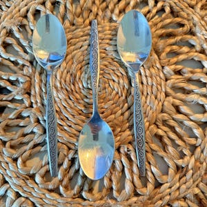 May include: Three silver spoons with ornate designs on the handles. The spoons are arranged in a triangular pattern on a woven straw mat.