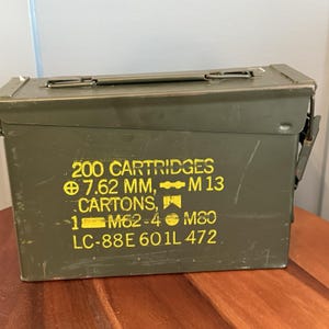 May include: An olive green metal ammunition box with a hinged lid and a latch closure. The box has yellow stenciled text that reads "200 CARTRIDGES 7.62 MM, M 13 CARTONS, 1 M62-4 M80 LC-88E 601L 472."