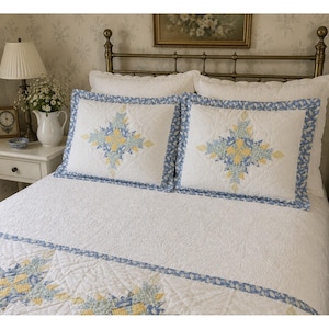 May include: A white quilt and two matching pillow shams featuring a central star pattern in yellow and blue floral fabric. The bedding set has a white quilted texture with blue floral borders and is displayed on a bed with a brass headboard.