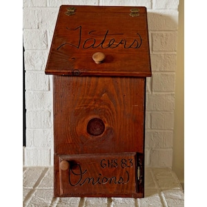 May include: A wooden storage box with a hinged lid and a small wooden knob. The lid is labeled "Taters" in cursive script. The lower section is labeled "Onions" and "GHS 83". The box is stained a warm brown color.