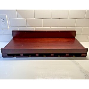 May include: A dark cherry-stained wooden wine glass rack. The rack has a shelf on top and slots underneath to hold wine glasses. The rack is designed to be mounted on a wall. The wood has a smooth finish.