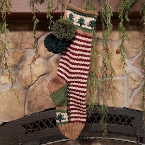 May include: A handmade Christmas stocking with a knitted design. The stocking features alternating burgundy and white stripes, with green and brown accents. It is adorned with green pom-poms and tree patterns. The stocking is hanging above a fireplace.