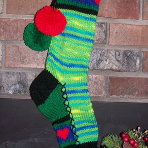 May include: A green, blue, and black striped Christmas stocking with red hearts and pom-poms. The stocking is hanging on a brick fireplace.